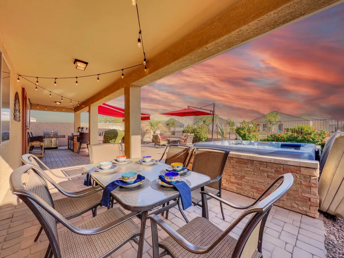 A beautifully set outdoor dining table under decorative lights, with a jacuzzi visible in the background and colorful umbrellas adding vibrancy. The scene captures a warm, inviting atmosphere perfect for evening meals, entertaining guests, or simply relaxing outdoors.