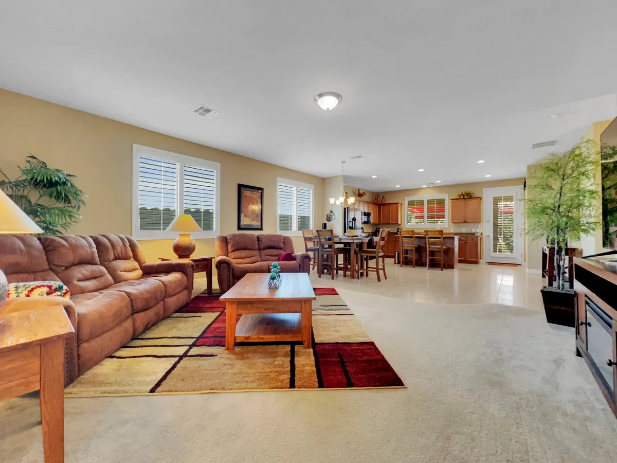 A wide shot showcasing the open-concept living area, featuring comfortable brown sofas, a decorative area rug, a wooden coffee table, a dining table and chairs, and the kitchen area in the background. The space is filled with natural light and set up for relaxation and social gatherings.