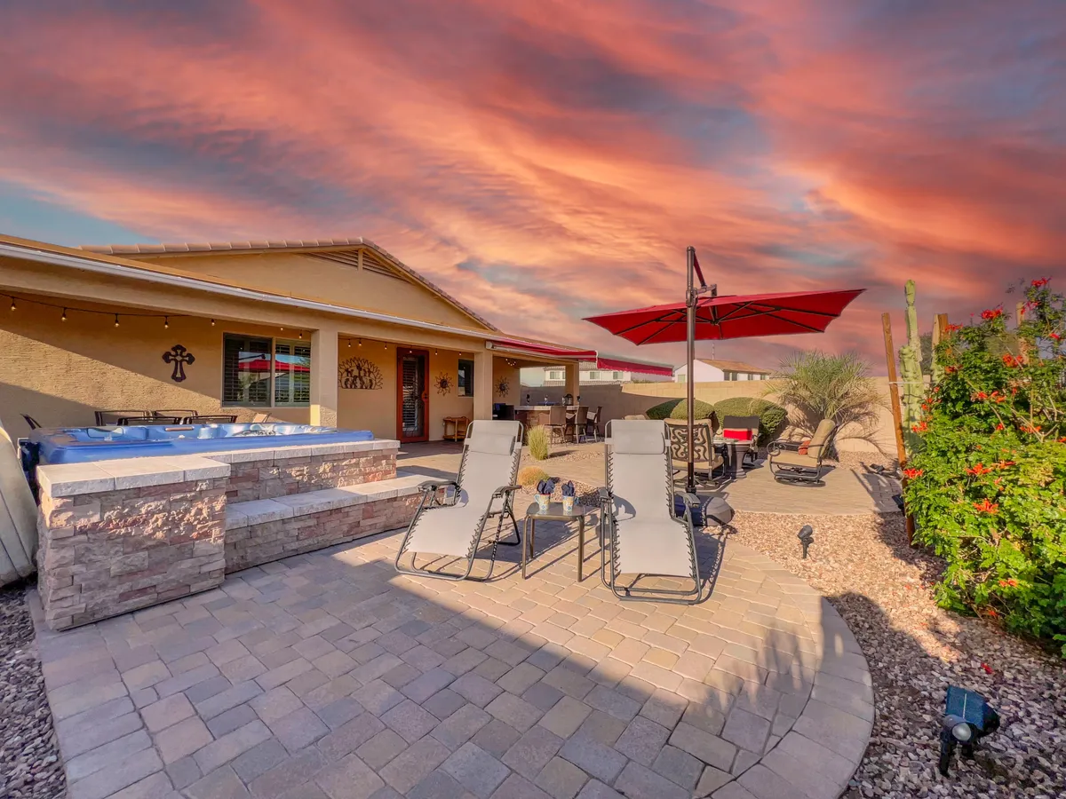 This exterior image showcases a beautifully landscaped backyard with a hot tub, lounge chairs, a red umbrella, and lush garden surroundings. The setting is perfect for relaxing outdoors, enjoying sunsets, and social gatherings.