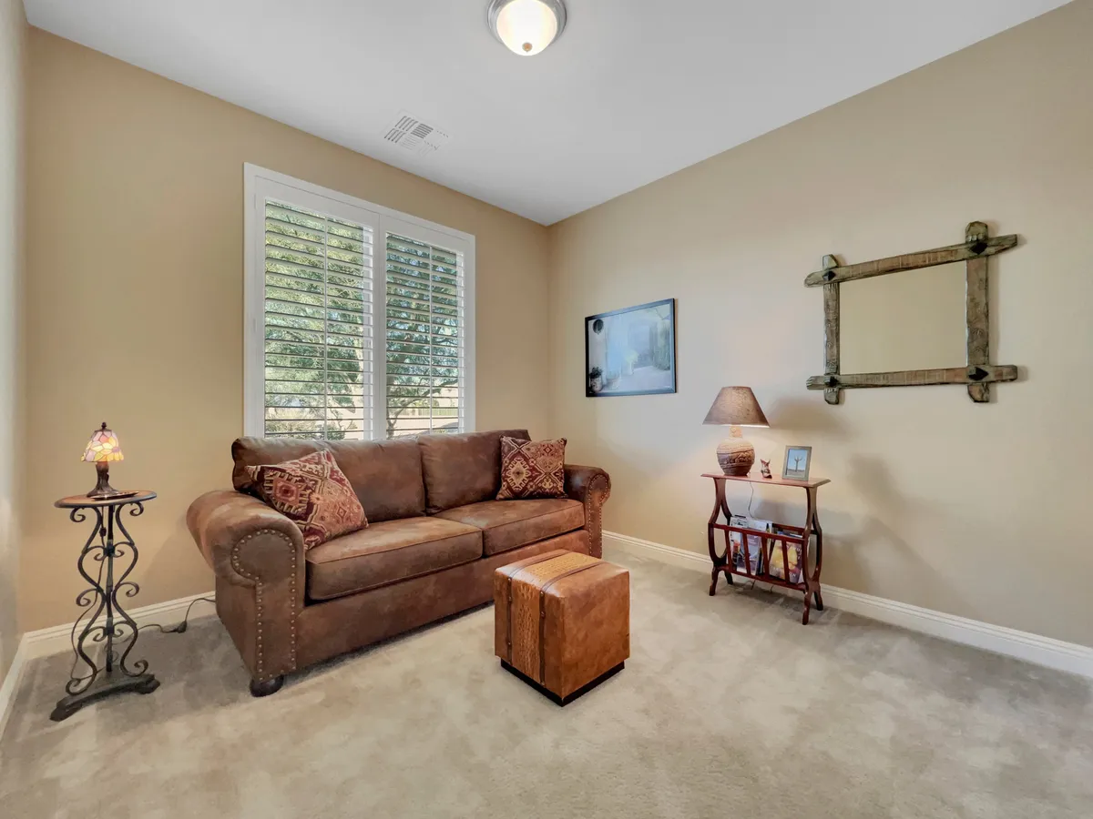 A cozy interior space with a brown sofa adorned with decorative pillows, a small wooden ottoman, a table lamp, and framed artwork on the wall. The setting invites relaxation, reading, or conversation, and exudes an inviting and homey atmosphere.