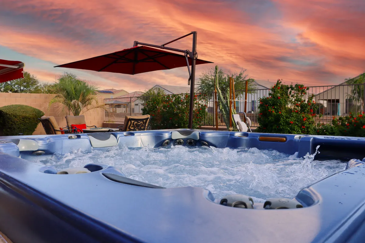This image features the raised hot tub as the focal point, surrounded by a well-landscaped garden and an inviting patio area. The scene is set at sunset, with warm golden light creating a serene and indulgent atmosphere. The presence of comfortable seating suggests space for conversation or relaxation, while the view extends to the desert landscape beyond, connecting the spa experience with nature.