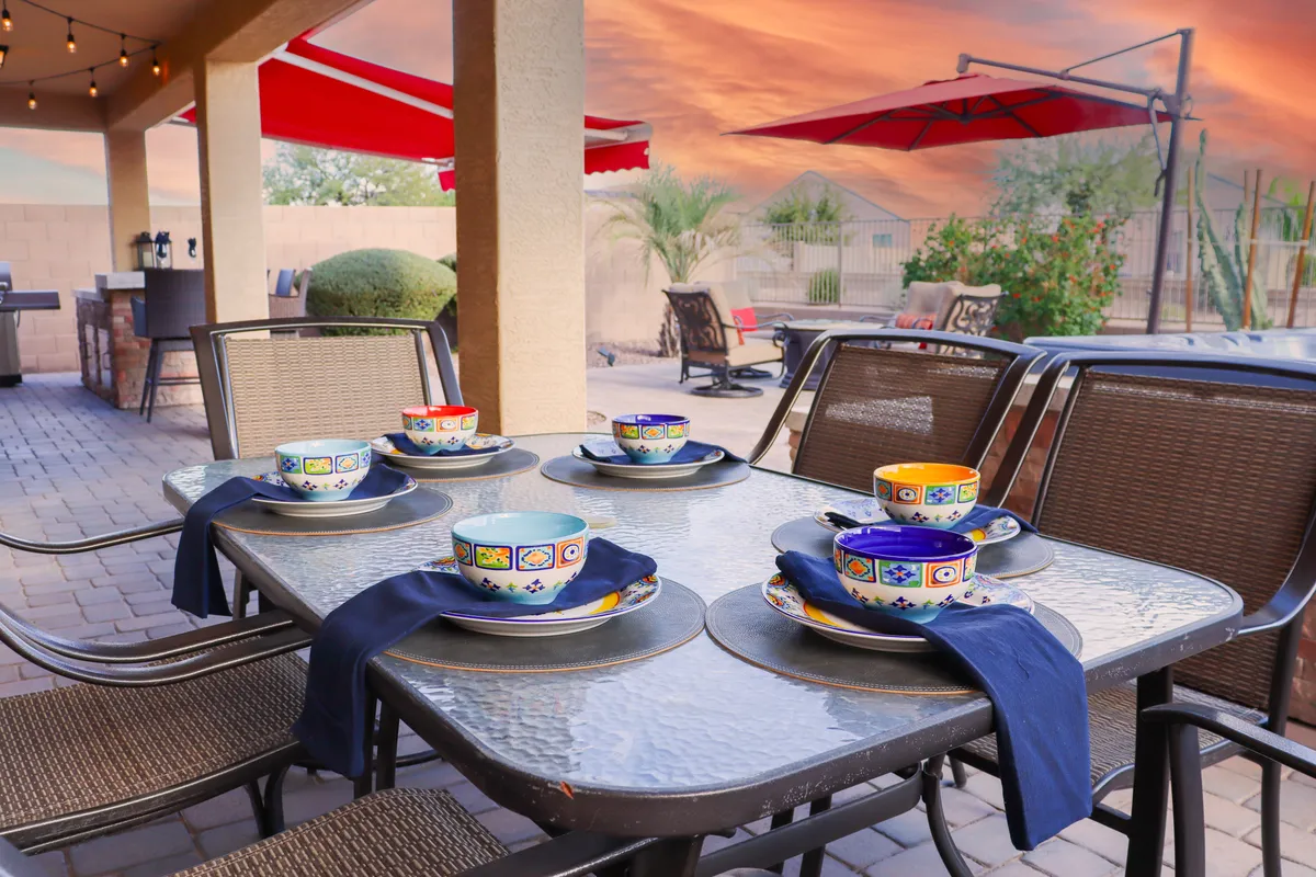 A vibrant outdoor dining area featuring a table set with colorful bowls, an outdoor kitchen, and cheerful red umbrellas. The setup is designed for group meals, celebrations, and social gatherings, all framed by an inviting patio atmosphere.